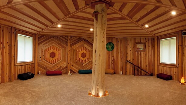 Een kamer met een hoge plafond, houten wanden en een centrale houten pilaar. Er zijn twee ramen in de wanden, een ingelijst kunstwerk en een klein tafeltje met een kaars.