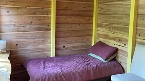 Une petite pièce chaleureuse aux murs en bois et au plafond en pente. On y trouve un lit recouvert d’une couverture violette et orné d’un seul oreiller, une petite table avec une lampe et une chaise.