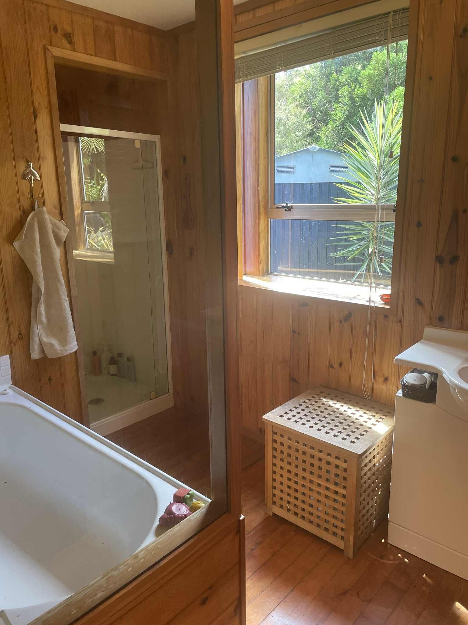 A wooden bathroom with a bathtub, a window with a view of greenery, and a laundry basket.