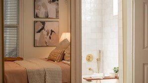 A bedroom with a bed, two wall-mounted abstract paintings, a bedside lamp, and a view into a bathroom with a bathtub and shower.