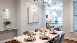 A dining area with a long table set for four, a chandelier, a mirror, and a framed abstract painting on the wall.