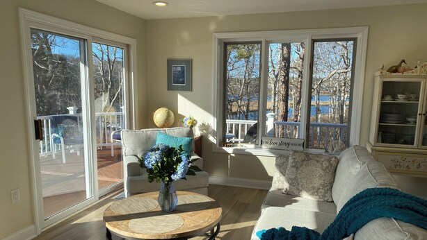 A living room with a sofa, a coffee table, and a view of a deck and trees. - New to the market 4BR Cape Cod charmer on Hamblin Pond w Waquoit Bay views (Mashpee)