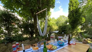 Un espacio al aire libre para comer con una mesa de madera dispuesta con comida y bebidas, rodeado de follaje y una hamaca.
