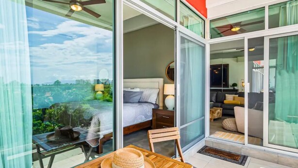 Une chambre moderne avec un lit, une petite table sur laquelle est posé un chapeau, et une vue sur le paysage urbain à travers de grandes portes vitrées.