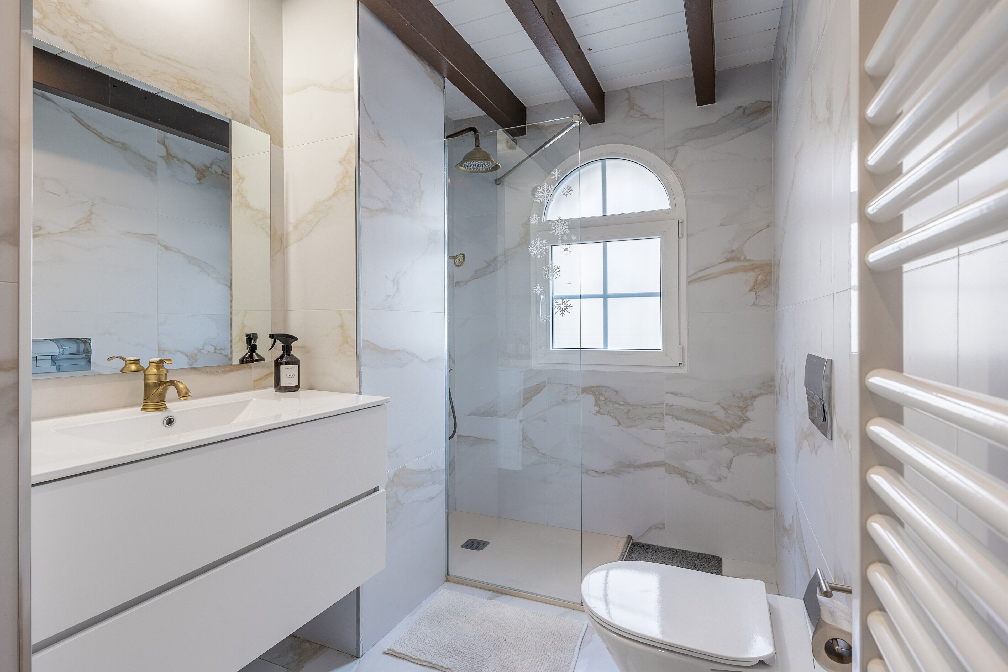 Un bagno con parete in marmo, box doccia in vetro, lavabo con rubinetteria dorata e una finestra.