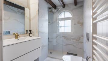 Un bagno con parete in marmo, box doccia in vetro, lavabo con rubinetteria dorata e una finestra.