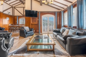 A spacious living room with a glass coffee table, leather sofas, wooden flooring, a dining area, and a large window with a view.