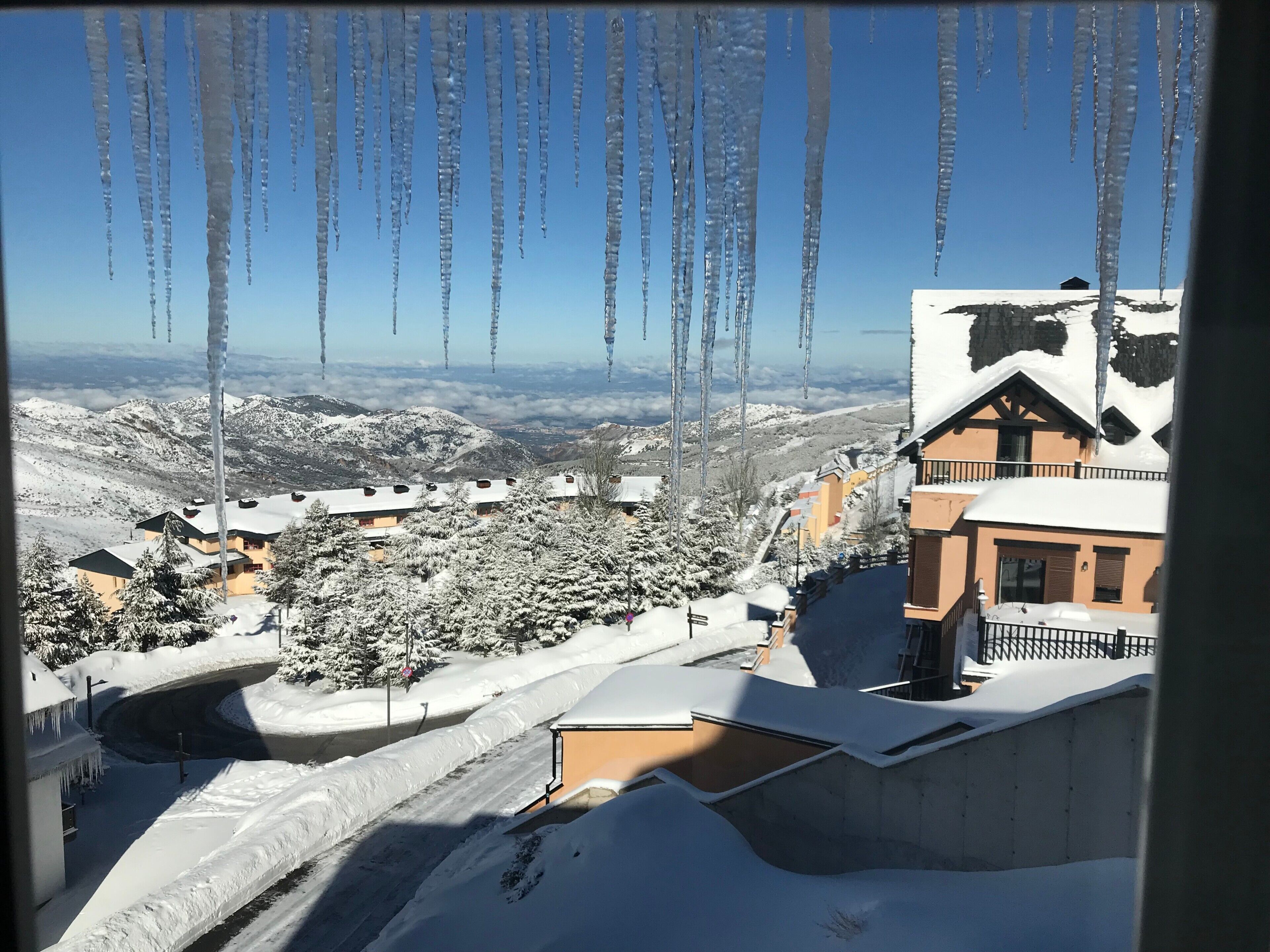 A snow-covered landscape with icicles hanging from a roof, a clear blue sky, and a view of a valley with buildings.