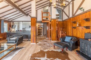 A spacious living area with a high ceiling, wooden beams, a staircase, leather furniture, and a kitchen in the background.