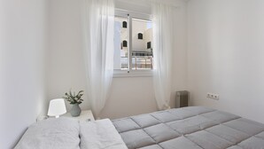 Un dormitorio con cama, una mesita de noche con lámpara, un florero con flores y una ventana con cortinas.