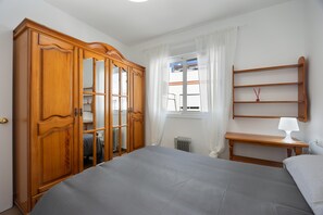 Un dormitorio con cama, un armario de madera, una mesita de noche con lámpara, ventana con cortinas y radiador.