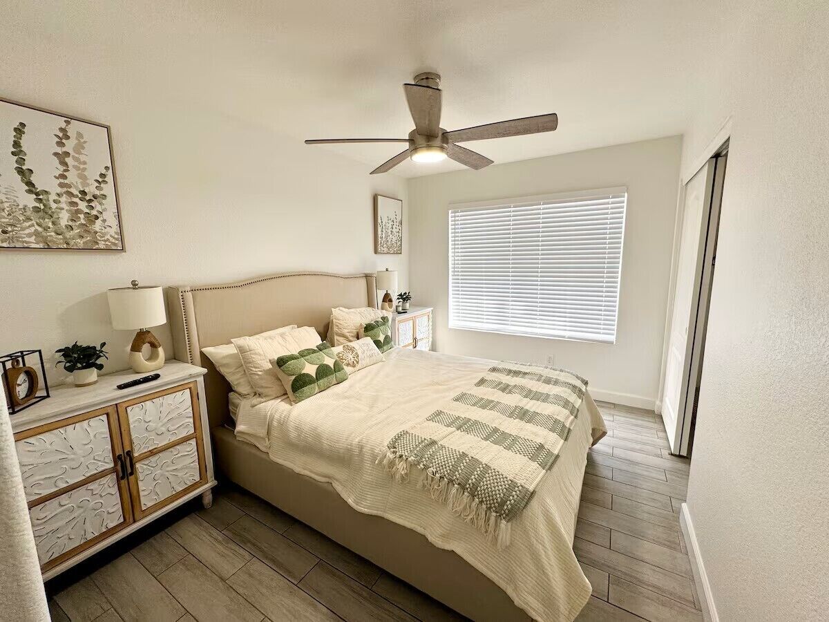 A bedroom with a bed, a nightstand, a ceiling fan, a window with blinds, and a wall-mounted artwork.