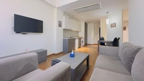 A modern living room with a flat-screen TV, grey sofas, a coffee table, and a kitchen in the background.