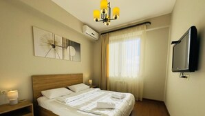 A neatly made bed with white linens, a wooden headboard, a bedside table with a lamp, and a wall-mounted TV.