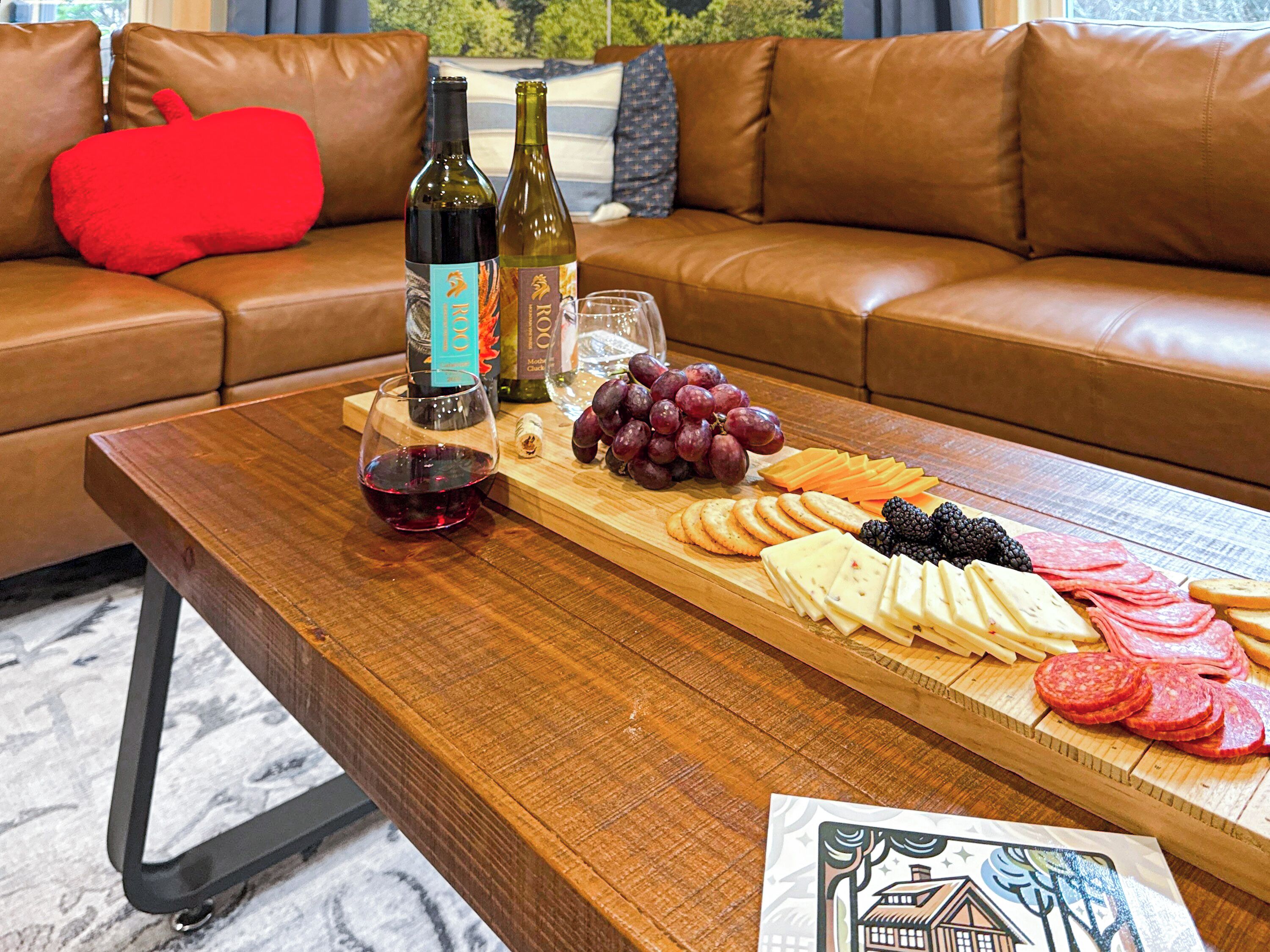 A wooden table with a wine bottle, a glass, and a cheese board with various cheeses, grapes, and crackers.