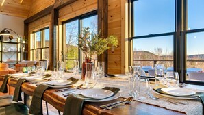 A wooden dining table set for a meal with clear glassware and plates, large windows with a view of the outdoors, and a vase with greenery.