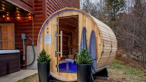 A wooden sauna with a door open, revealing a bench inside. There are two potted plants outside the sauna.