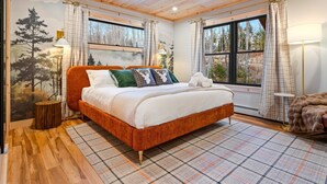 A bedroom with a wooden ceiling, a bed with white bedding, plaid curtains, and a view of trees through the window.