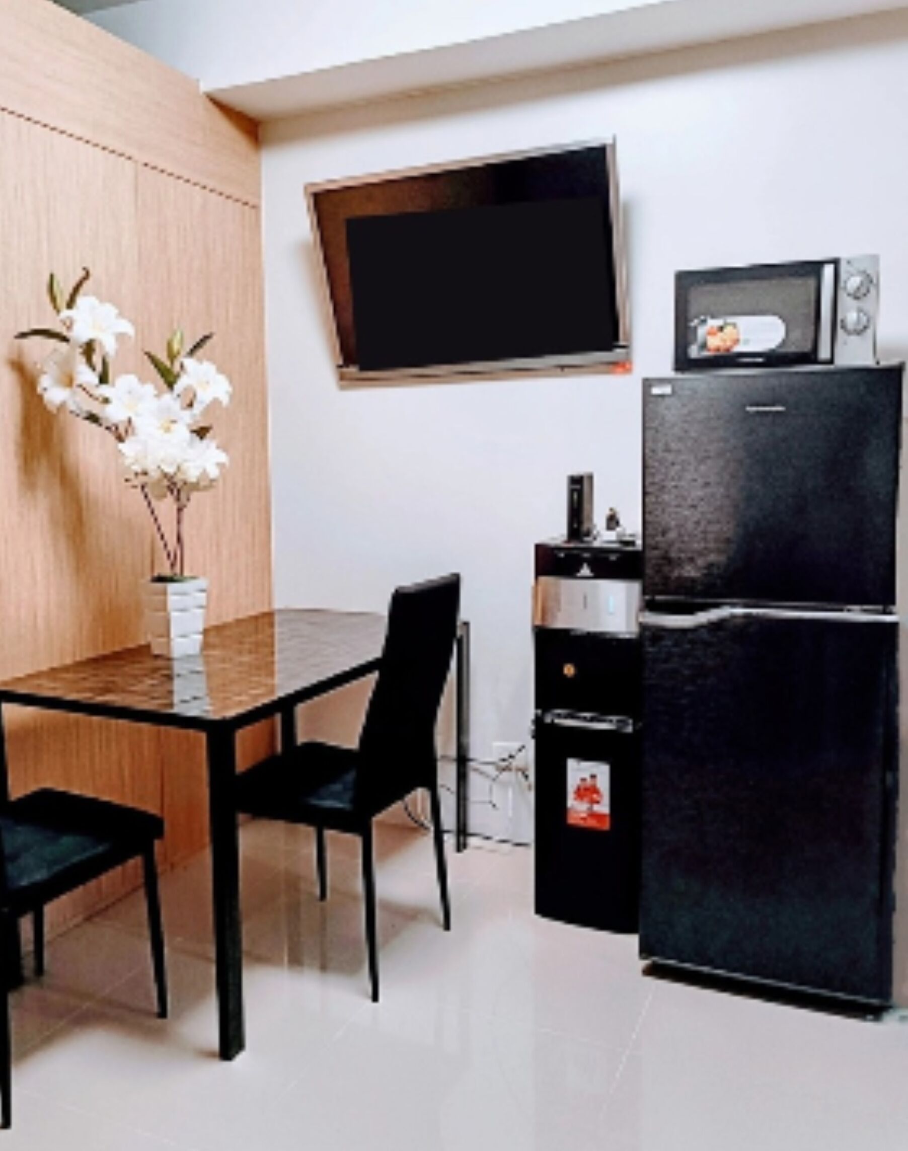 Une cuisine de taille réduite avec une table en bois, des chaises noires, un réfrigérateur noir, un four à micro-ondes et un téléviseur à écran plat fixé au mur.