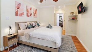 Une chambre à coucher avec un lit, un ventilateur de plafond, des œuvres d’art accrochées au mur, une télévision et une table de chevet.