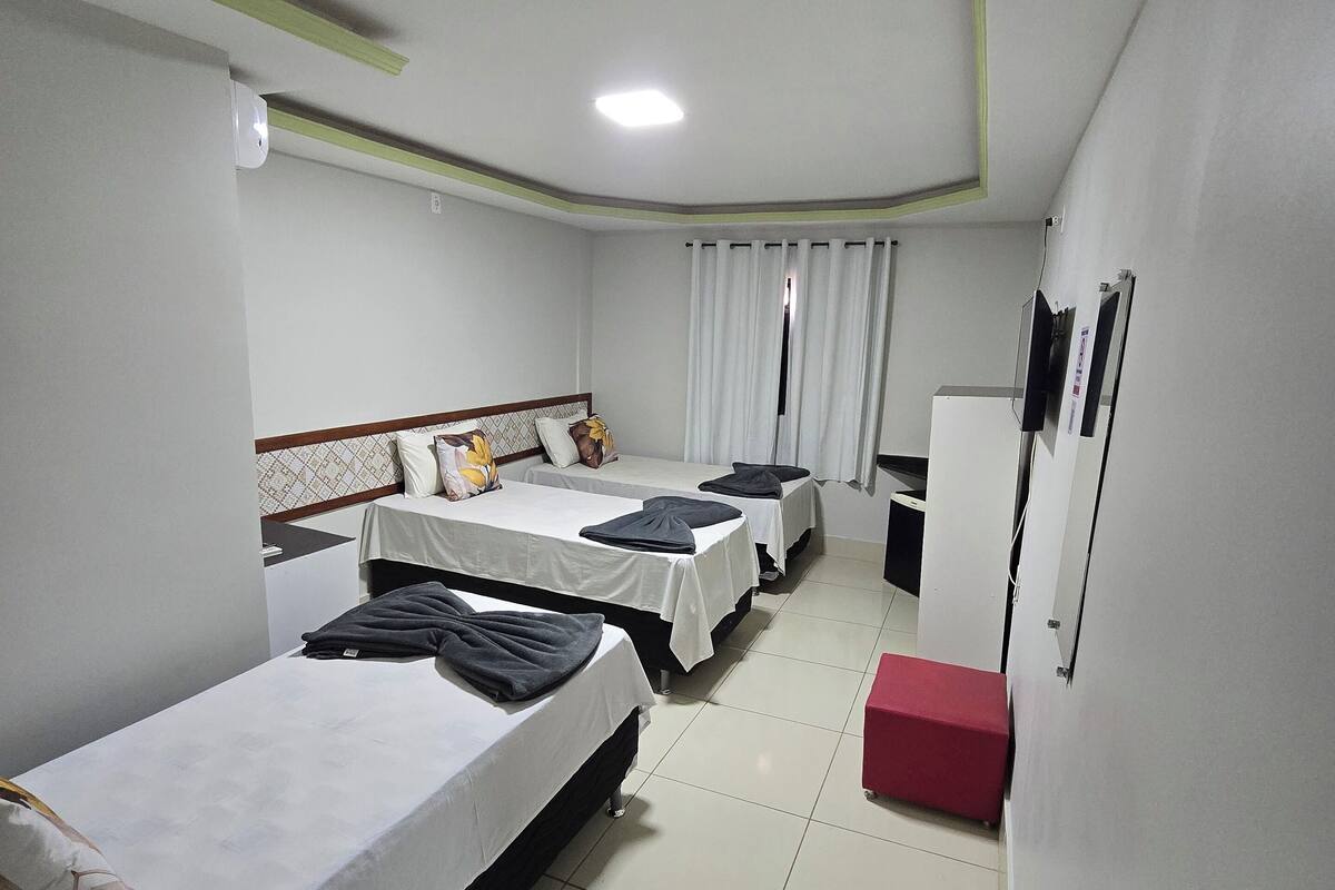 A hotel room with three single beds, a small red bench, a television, and a window with curtains.