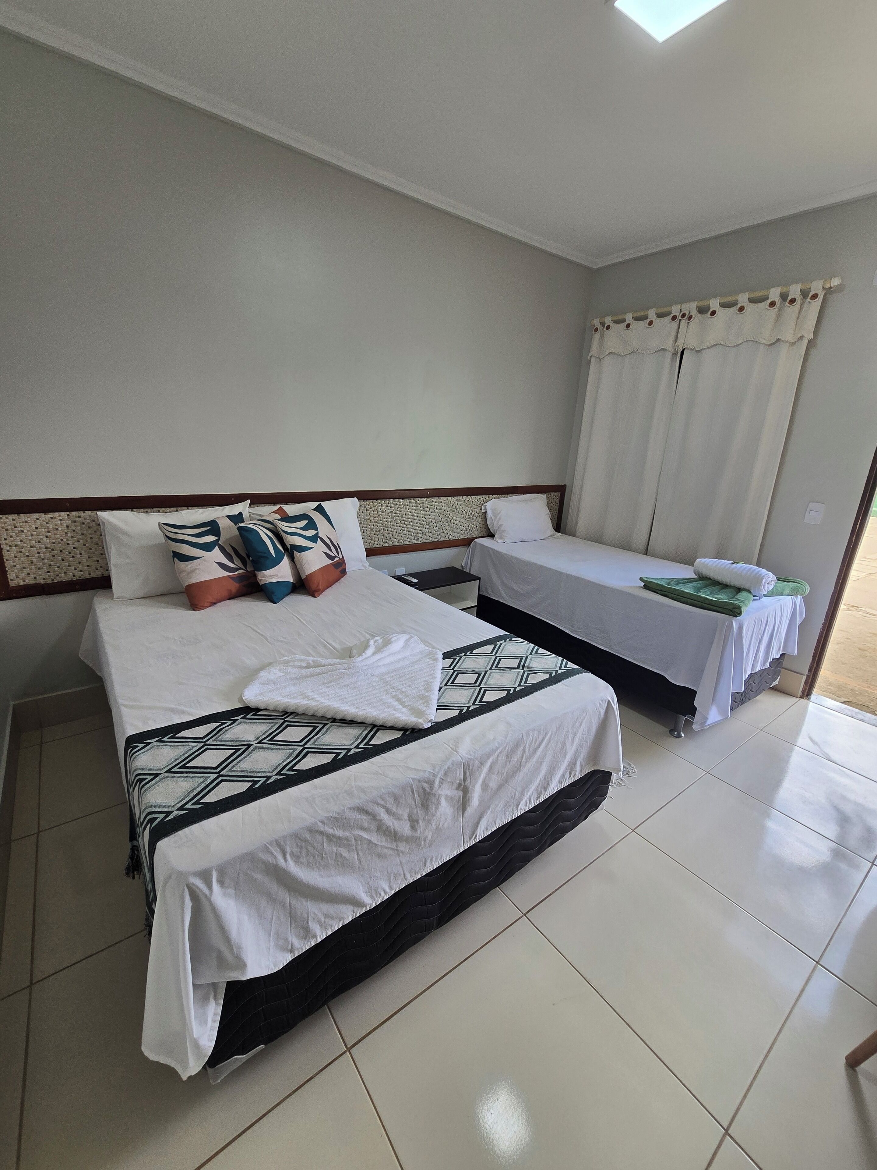 A hotel room with two beds, a tiled floor, and a window with curtains.