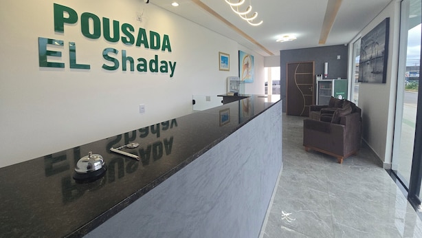 A reception area with a marble counter, a sign reading "POUSADA EL Shaday," and a seating area with a sofa and chairs. - Pousada Elshaday (Vitória da Conquista)