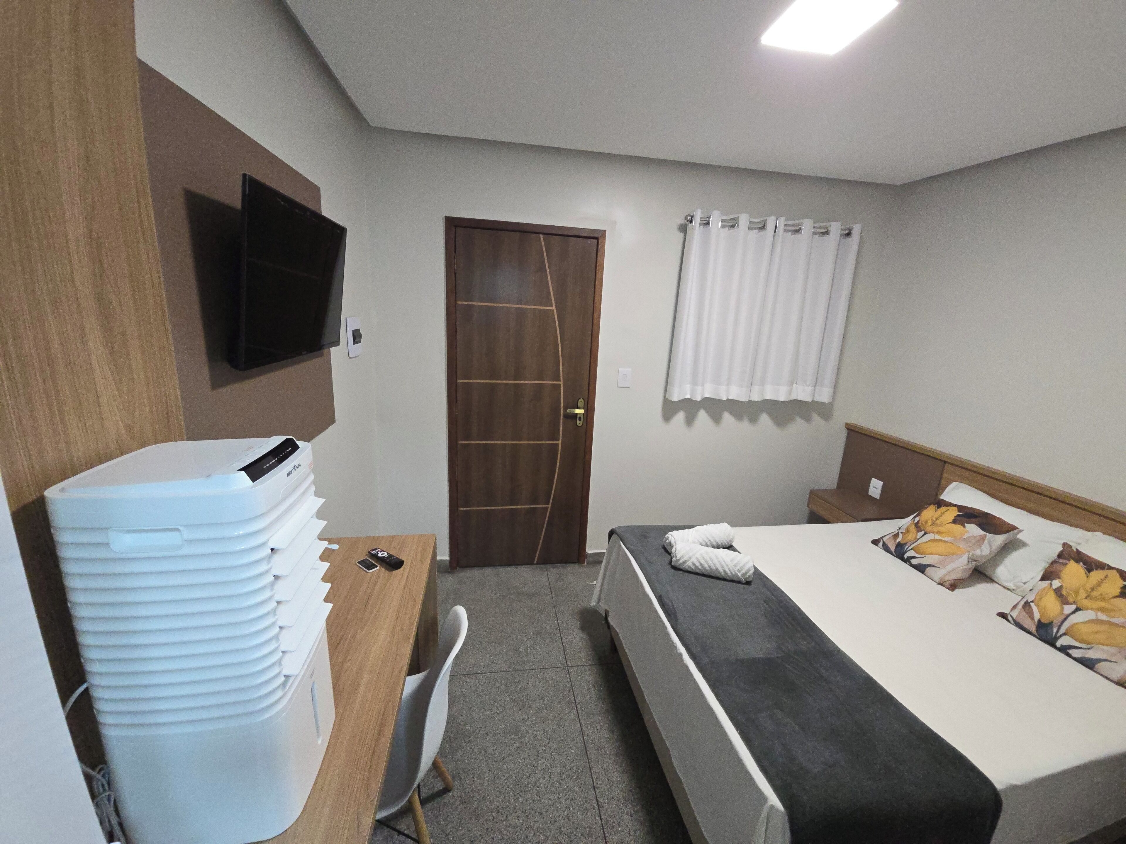 A hotel room with a bed, a desk with a computer, a chair, a TV mounted on the wall, and a door with a curtain.