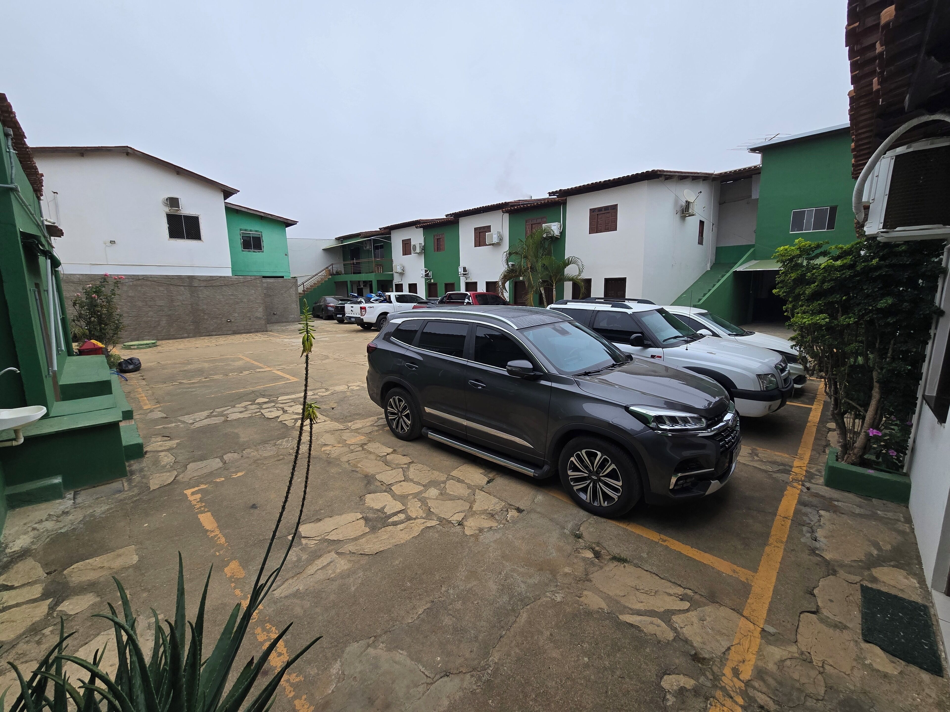 Uma área de estacionamento com vários carros, edifícios verdes e brancos e um chão pavimentado em pedra.