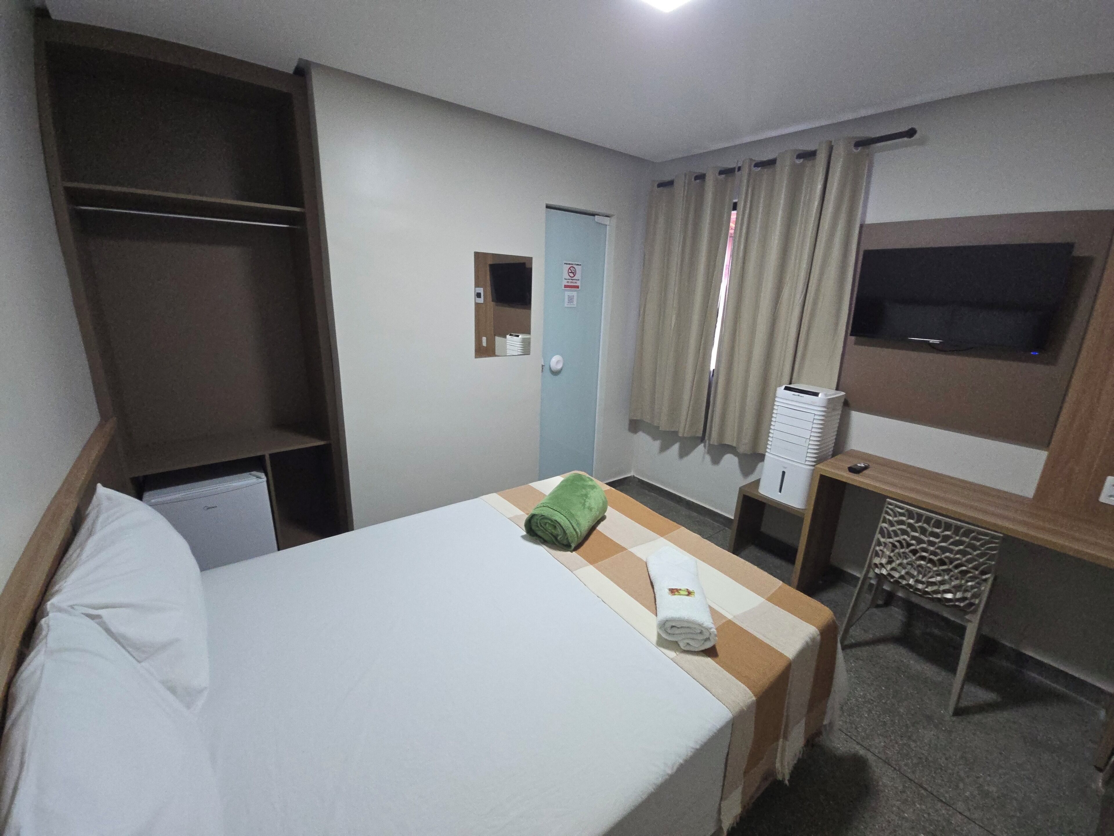 Quarto de hotel com cama, escrivaninha, televisão e guarda-roupa.