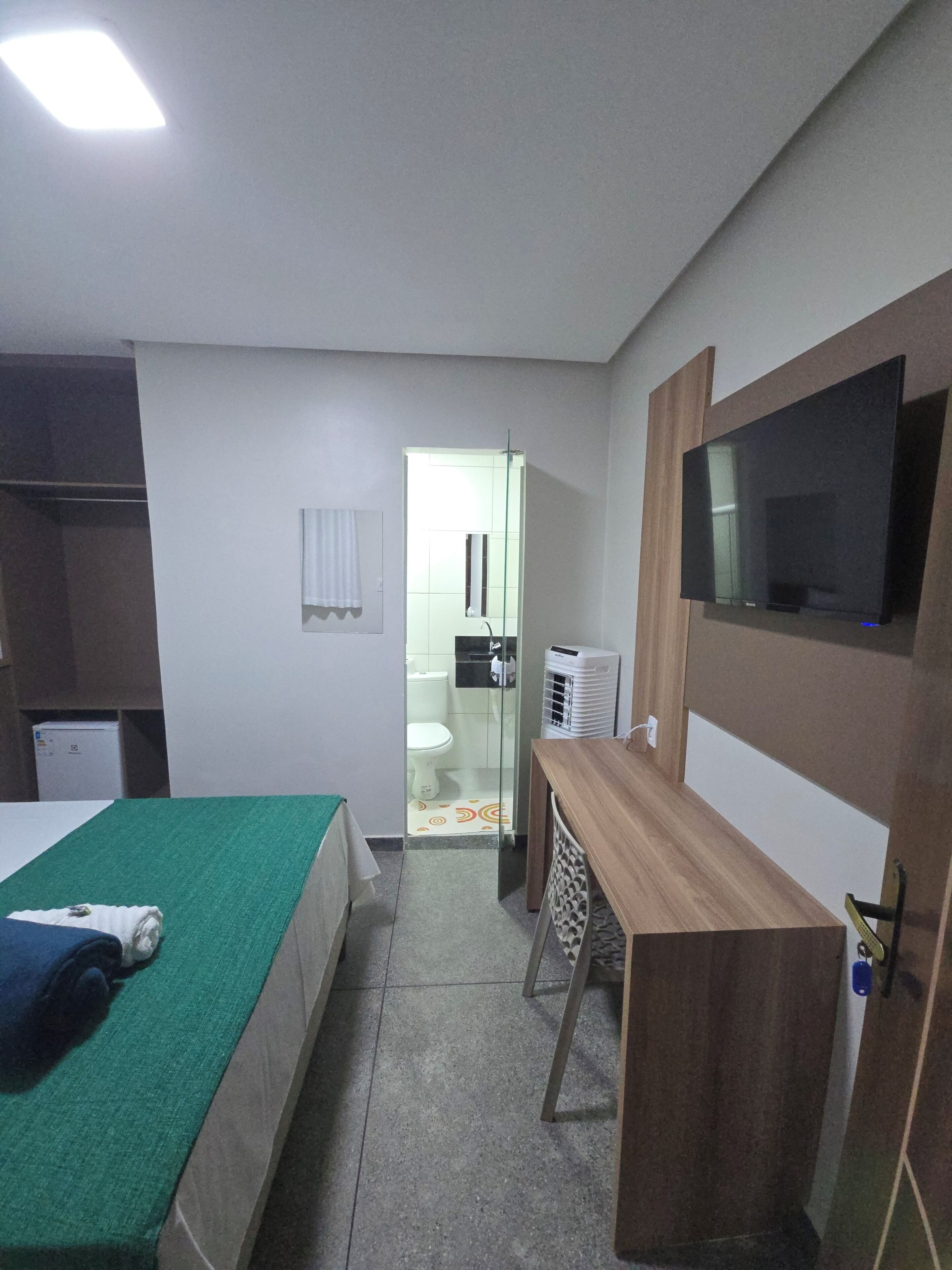 A hotel room with a bed, a desk, a TV, a wardrobe, and a bathroom visible through a glass door.