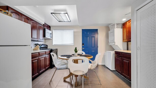 A compact kitchen with wooden cabinets, a white refrigerator, a blue door, and a dining area with white chairs and a round table. - Brighton Retreat - Walk to Metro (Seattle)