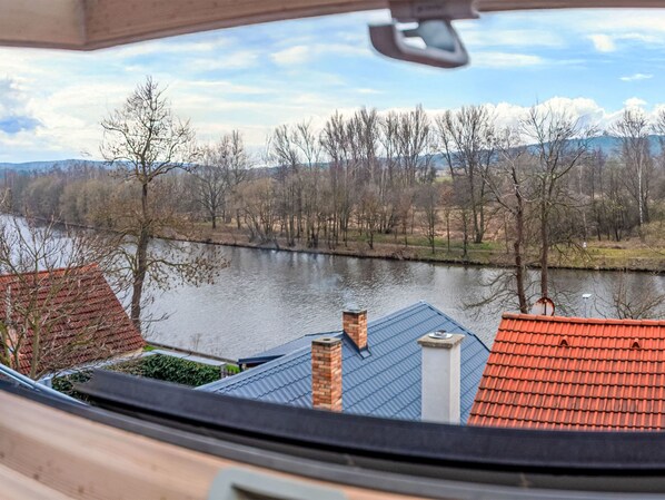 Un río con árboles y montañas al fondo, visto desde una ventana con tejados rojos.