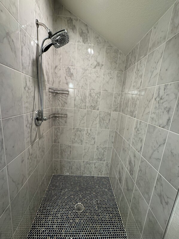 Een douche met een handdouche en een kraan aan de muur, een metalen douchebak, en een betegelde wand met een marmerachtig patroon.