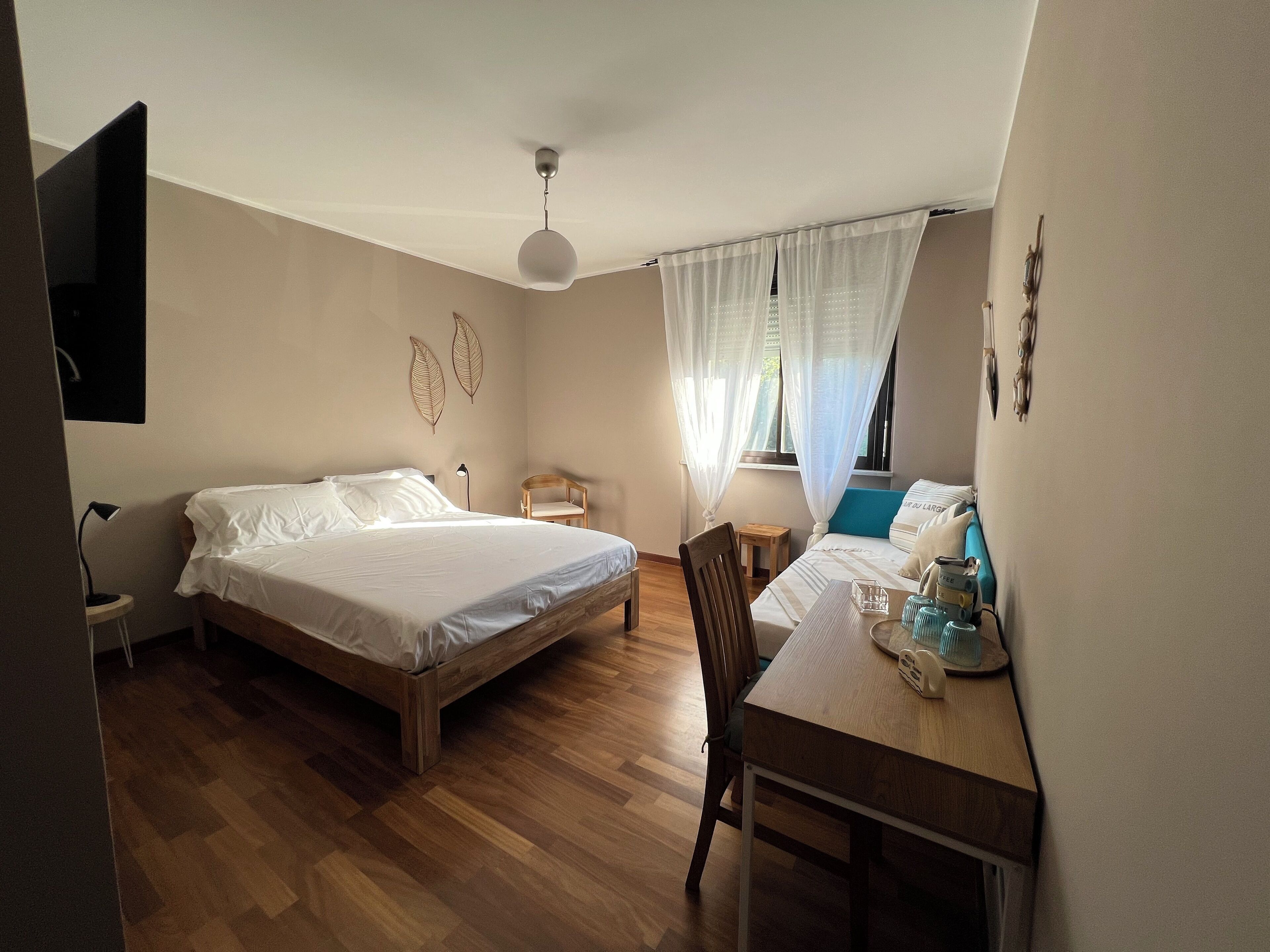 A bedroom with a wooden bed, a desk with a chair, a small table, and a window with sheer curtains.