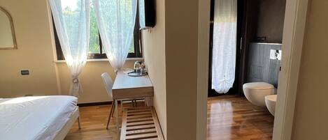 Un dormitorio con una cama, un escritorio y una silla. Se observa una ventana con cortinas translúcidas y vistas al verdor exterior.