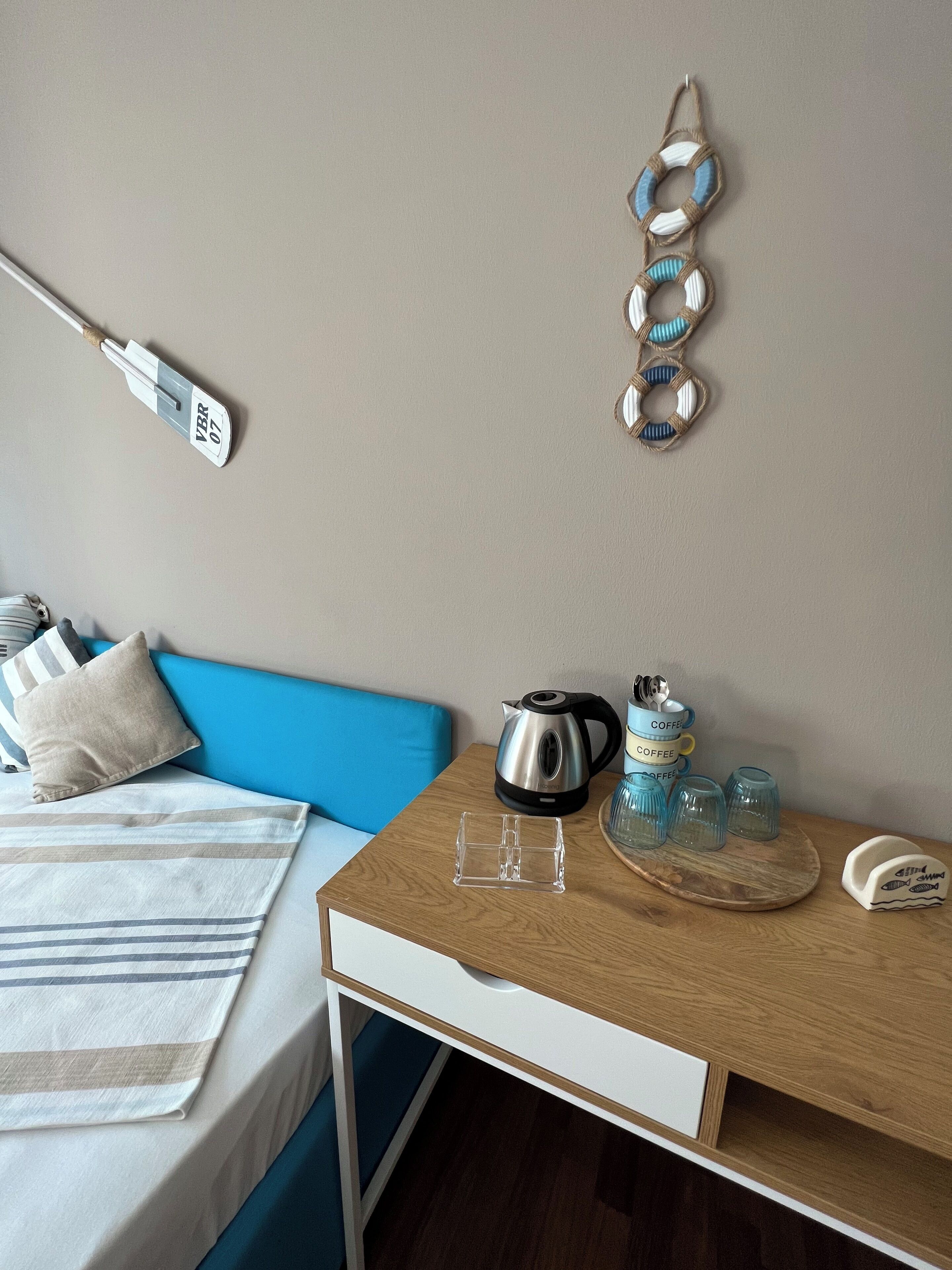 A neatly arranged hotel room with a bed, a blue headboard, a wooden side table with a kettle, coffee cups, and glasses.