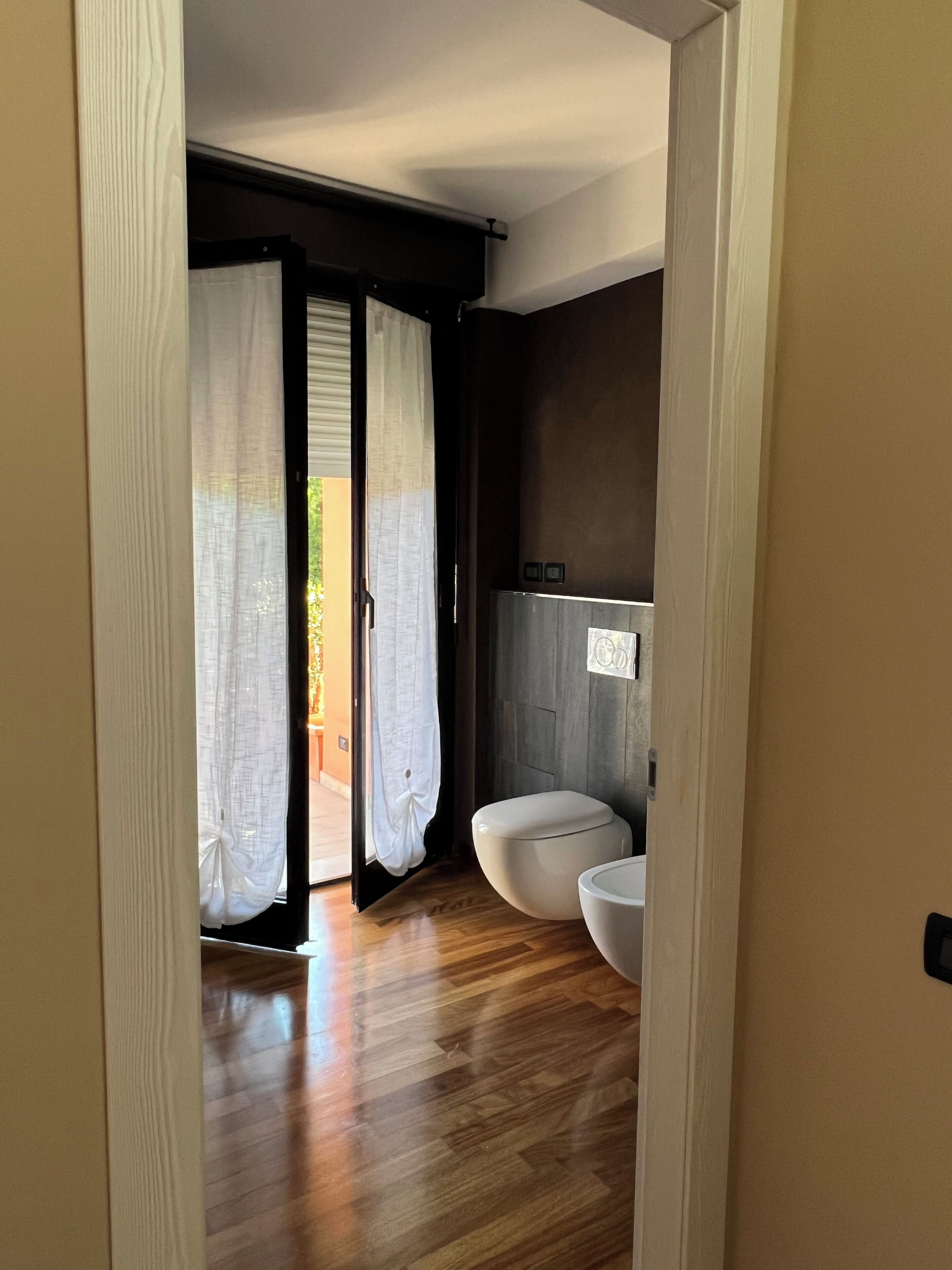 A modern bathroom with a toilet and a sink, wooden flooring, and a view of the outdoors through a door with sheer curtains.