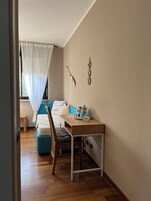 Un dormitorio con cama, mesita de noche, silla, ventana con cortinas traslúcidas y lámpara de pared.