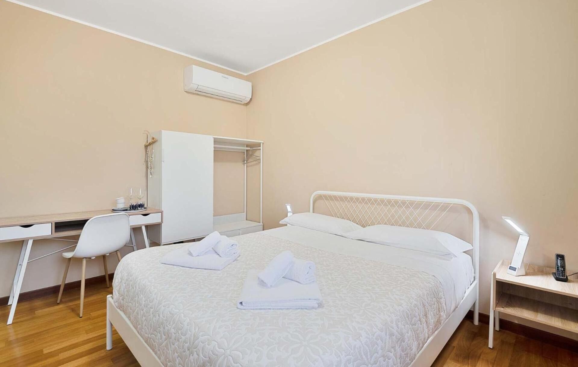 A hotel room with a bed, a desk, a chair, a wardrobe, and an air conditioning unit.