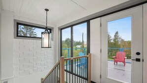 A room with a white wall, a hanging lantern, a wooden staircase, and a sliding glass door leading to an outdoor area with a red chair and blue deck. - Modern PEI Shipping Container Retreat with Multiple Decks & Stunning Views (Oyster Bed Bridge)