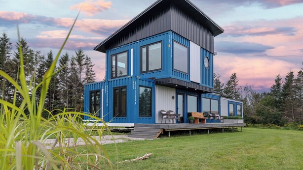 A modern multi-story house with a flat roof, large windows, and a deck with outdoor seating. - Modern PEI Shipping Container Retreat with Multiple Decks & Stunning Views (Oyster Bed Bridge)