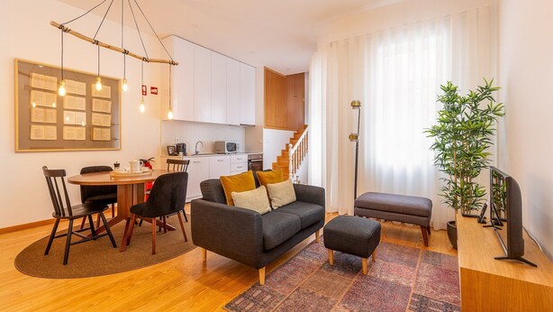 A modern living room with a sofa, ottoman, and coffee table, a dining area with chairs and a table, a kitchen area, and a staircase leading to an upper floor.