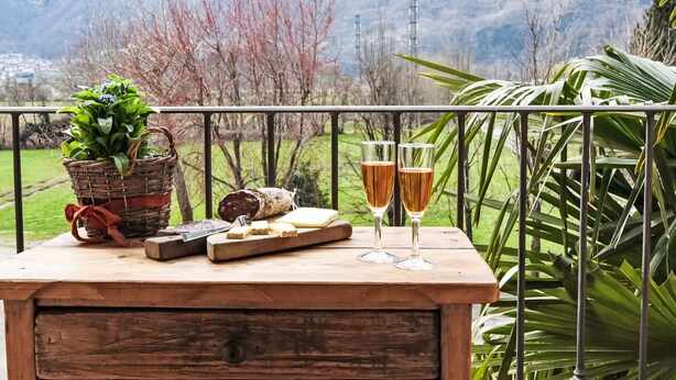 Una mesa de madera con una cesta de flores, una hogaza de pan, una tabla de queso y dos copas de vino en un balcón con barandilla de metal.