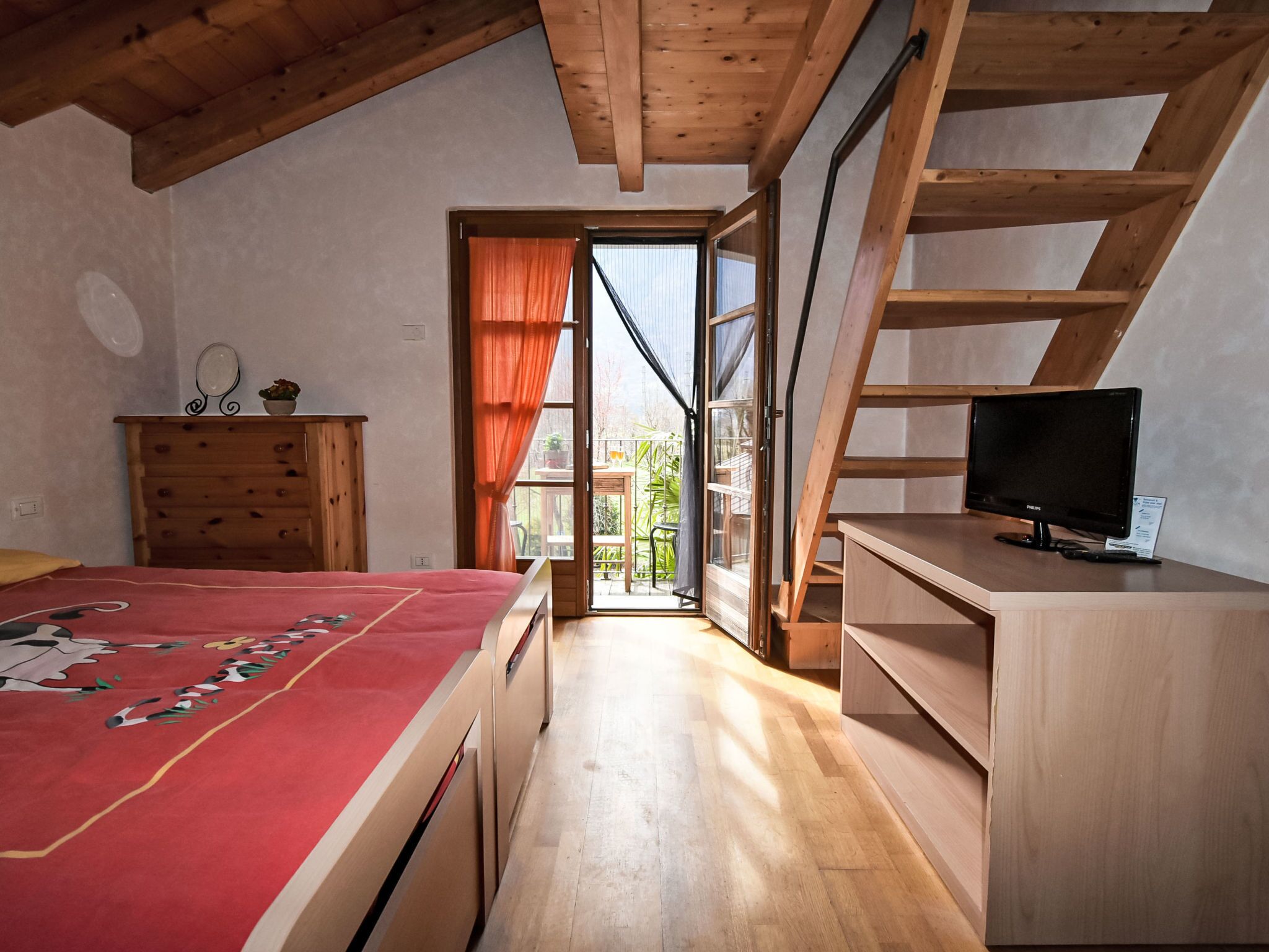 A bedroom with a bed, a dresser, a TV, and a window leading to a balcony.