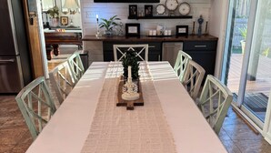 A dining area with a long table, chairs, and a decorative centerpiece. There is a kitchen in the background with a refrigerator, cabinets, and a countertop. - Welcome to our home! Lakefront Living, Where Beautiful Sunsets are unforgettable (Lake Park)
