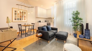 A modern living room with a sofa, ottoman, and a dining area with a table and chairs. There is a kitchen in the background with white cabinets and a plant on the windowsill. - Well-equipped apartment in Oporto (Oporto)