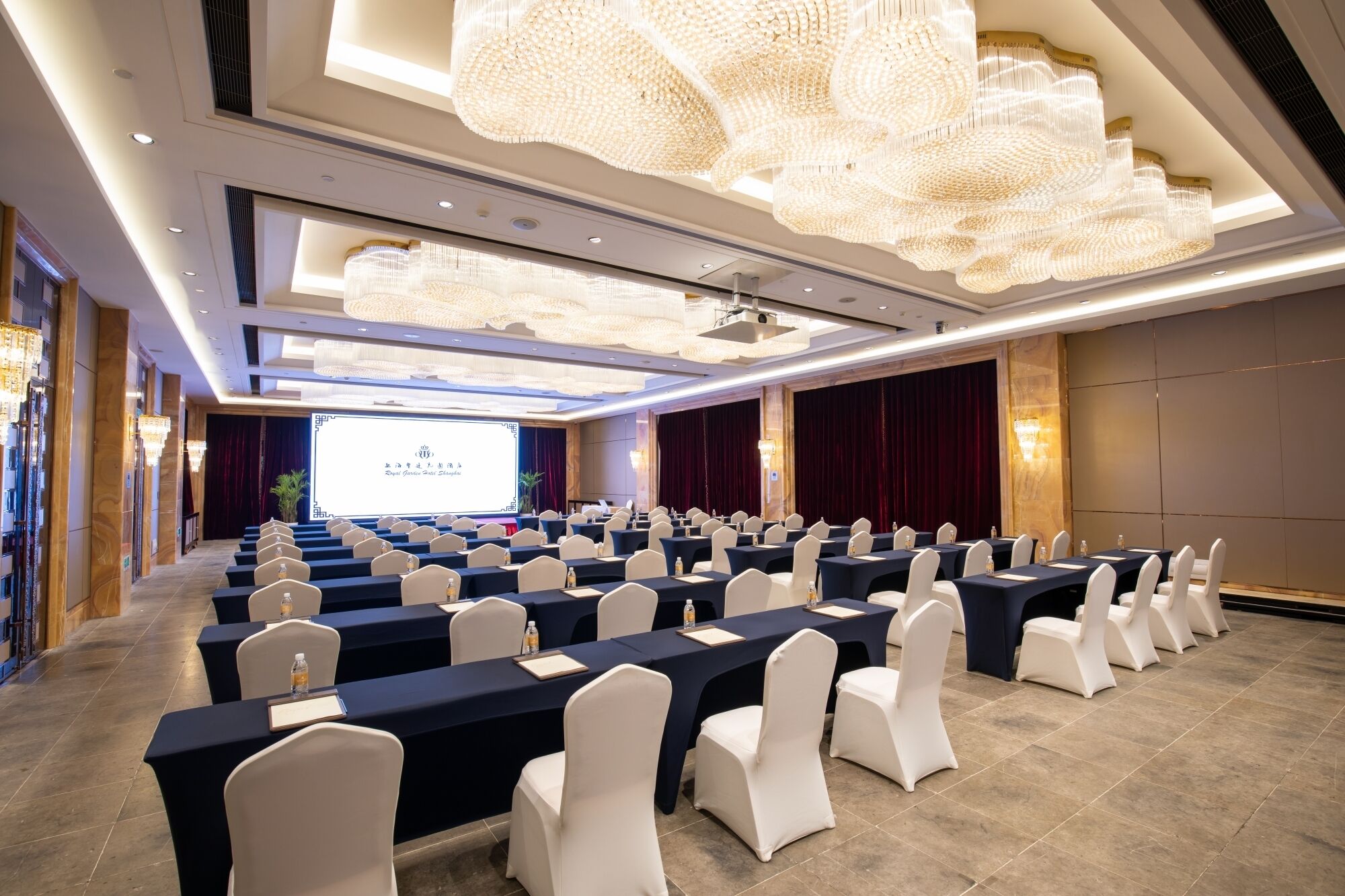 A conference room with rows of tables and chairs, a large screen at the front, and decorative chandeliers.