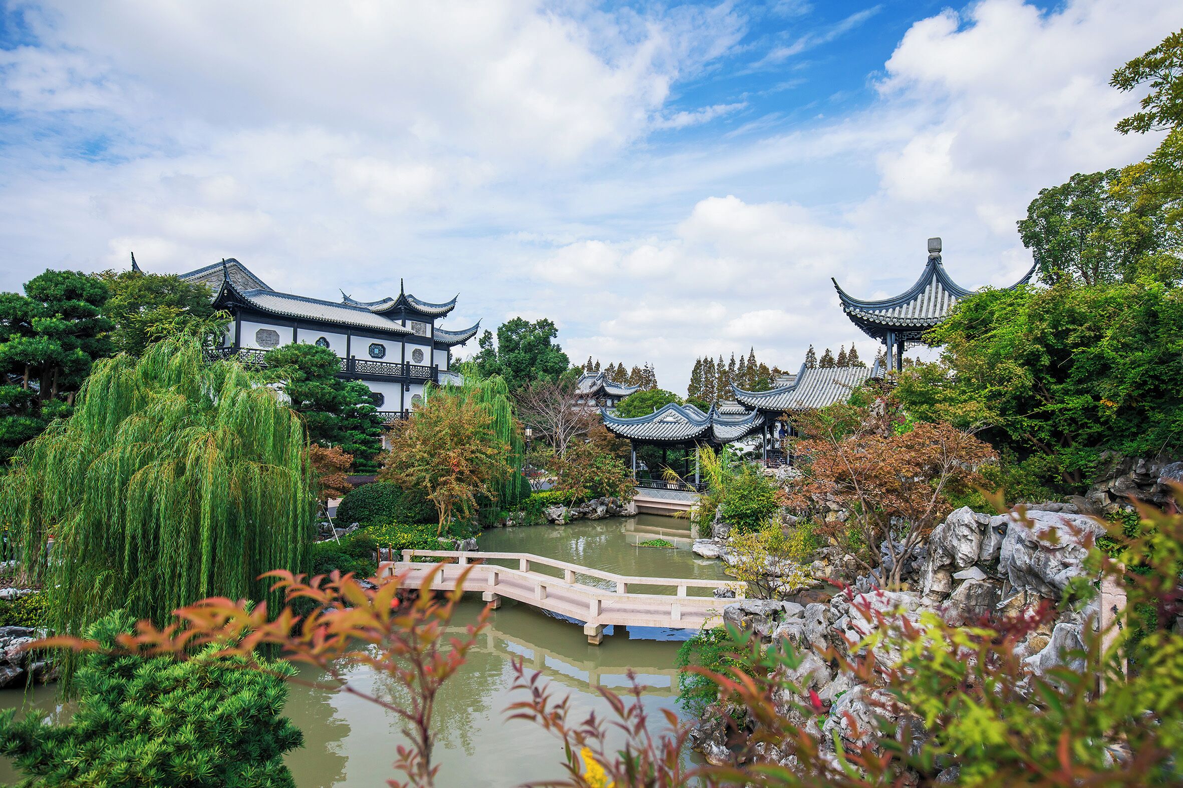 Un jardín chino tradicional con un puente de madera sobre un estanque, rodeado de abundante vegetación y arquitectura clásica.