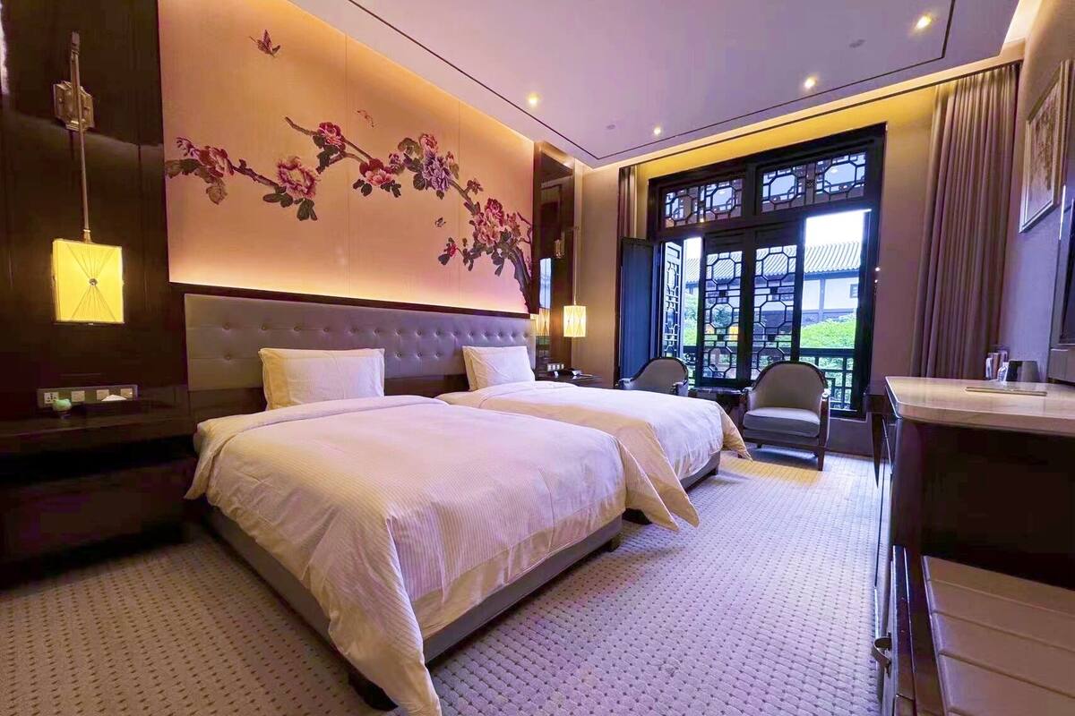 Une chambre d’hôtel avec un grand lit, deux tables de chevet, un bureau et une vue sur la verdure par la fenêtre.